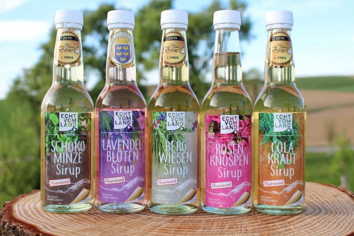 Fünf Flaschen Sirup sind auf einer Holzoberfläche ausgestellt. Sie haben weiße Kappen und sind mit verschiedenen Farben und Namen beschriftet. Von links nach rechts sind die Flaschen als Chokomintze Sirup, Lavendel Blueten Sirup, Berg Wiesen Sirup, Rosen Knospen Sirup und Cola Kraut Sirup gekennzeichnet.