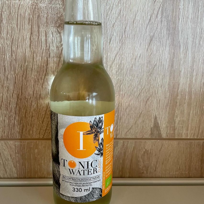Eine Flasche Tonic Water, 330 ml, auf einem hölzernen Hintergrund. Das Etikett zeigt einen orangefarbenen Kreis mit weißem Text und botanischen Illustrationen.