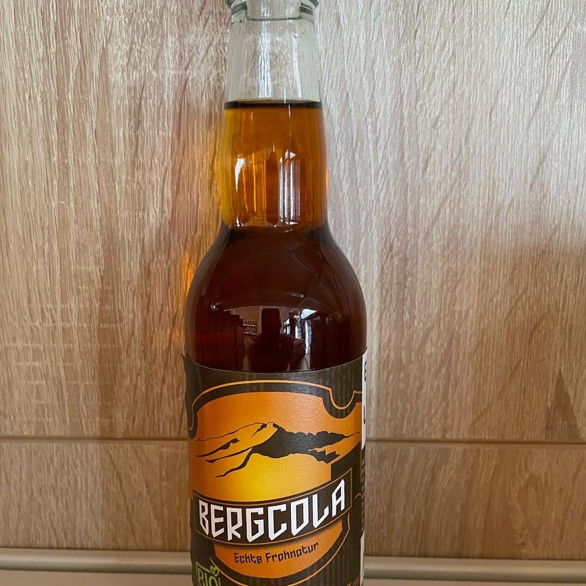 Eine Flasche Bier steht auf einer Holzoberfläche. Sie hat eine klare Glasflasche mit einer braunen Flüssigkeit darin. Das Etikett zeigt eine Bergkette und das Wort 'BERGCOLA'. Der Markenname 'Echte Frohntur' ist ebenfalls auf dem Etikett gedruckt.