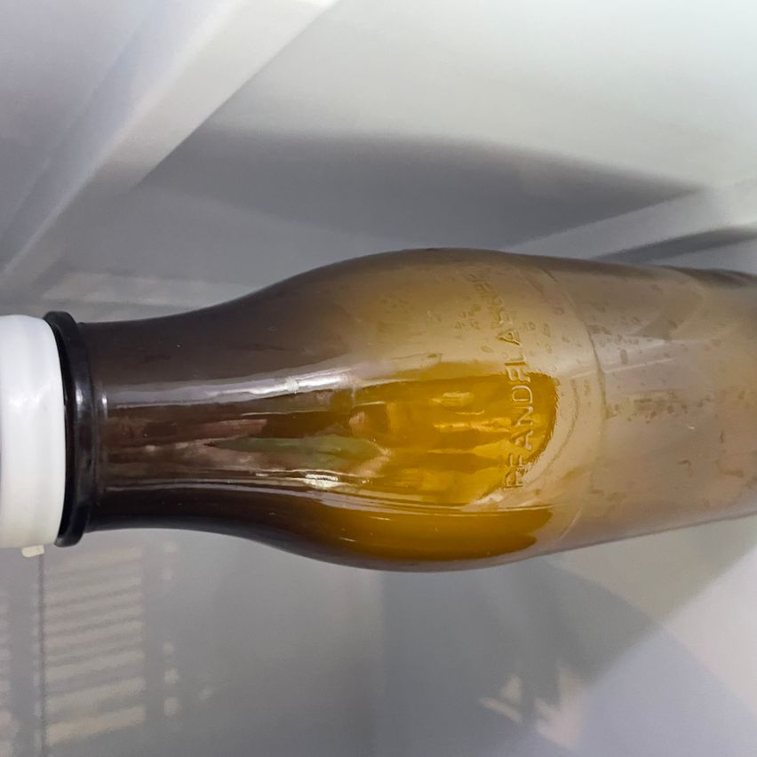 Eine Nahaufnahme einer teilweise gefüllten braunen Flasche mit weißem Verschluss, beschriftet mit 'PEANDE FLAVORED', auf einer weißen Oberfläche.