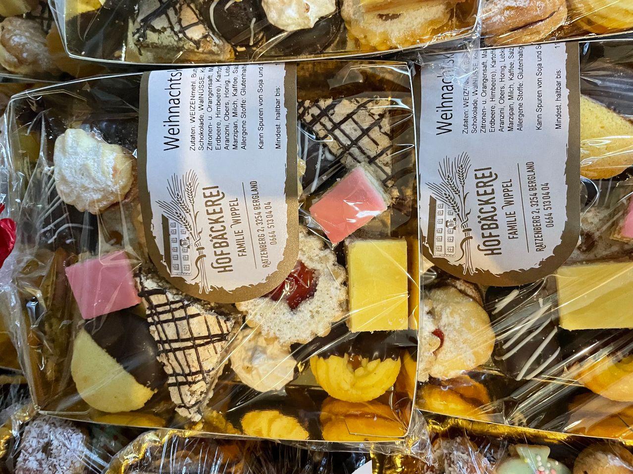Verpackte Packungen mit verschiedenen deutschen Weihnachtsplätzchen, einschließlich Schokoladenüberzogener und rosa-glasierter Gebäckstücke, sind in durchsichtige Plastikfolie eingewickelt. Die Etiketten kennzeichnen das Produkt als 'Weihnachts' und 'Hoebackerei'.