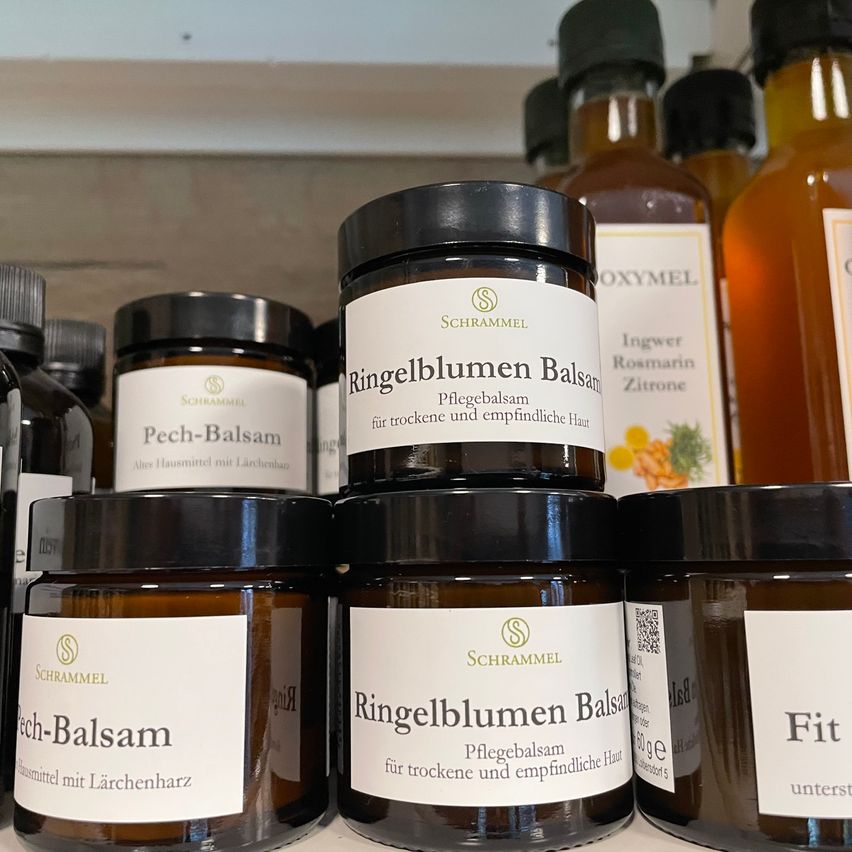 Verschiedene Gläser mit Ringelblumen Balsam stehen auf einem Regal. Sie sind für trockene und empfindliche Haut. Einige Etiketten enthalten 'Pflegebalsam'. Der Hintergrund ist unscharf.