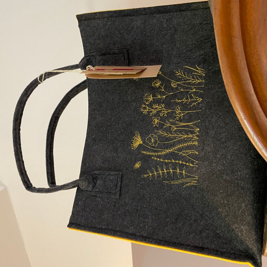 Eine graue Filztasche mit gelber Blumenstickerei auf der Vorderseite. Die Tasche hat zwei Henkel und einen Anhänger. Ein hölzerner Teller ist teilweise auf der rechten Seite sichtbar.
