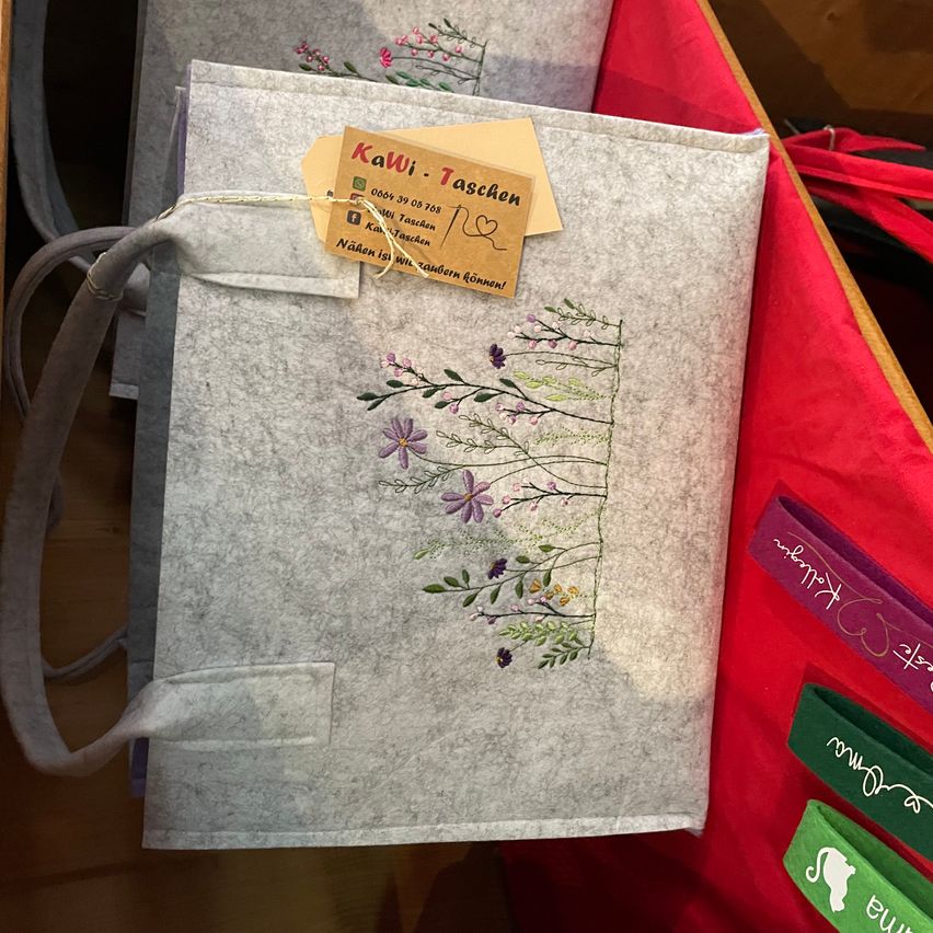 Eine graue filztasche mit Blumendesign auf einer roten Oberfläche. Ein Etikett mit 'Kawi Taschen' und einem Herzsymbol ist angebracht. Andere Gegenstände wie Bänder und ein katzenthematisches Objekt sind in der Nähe.