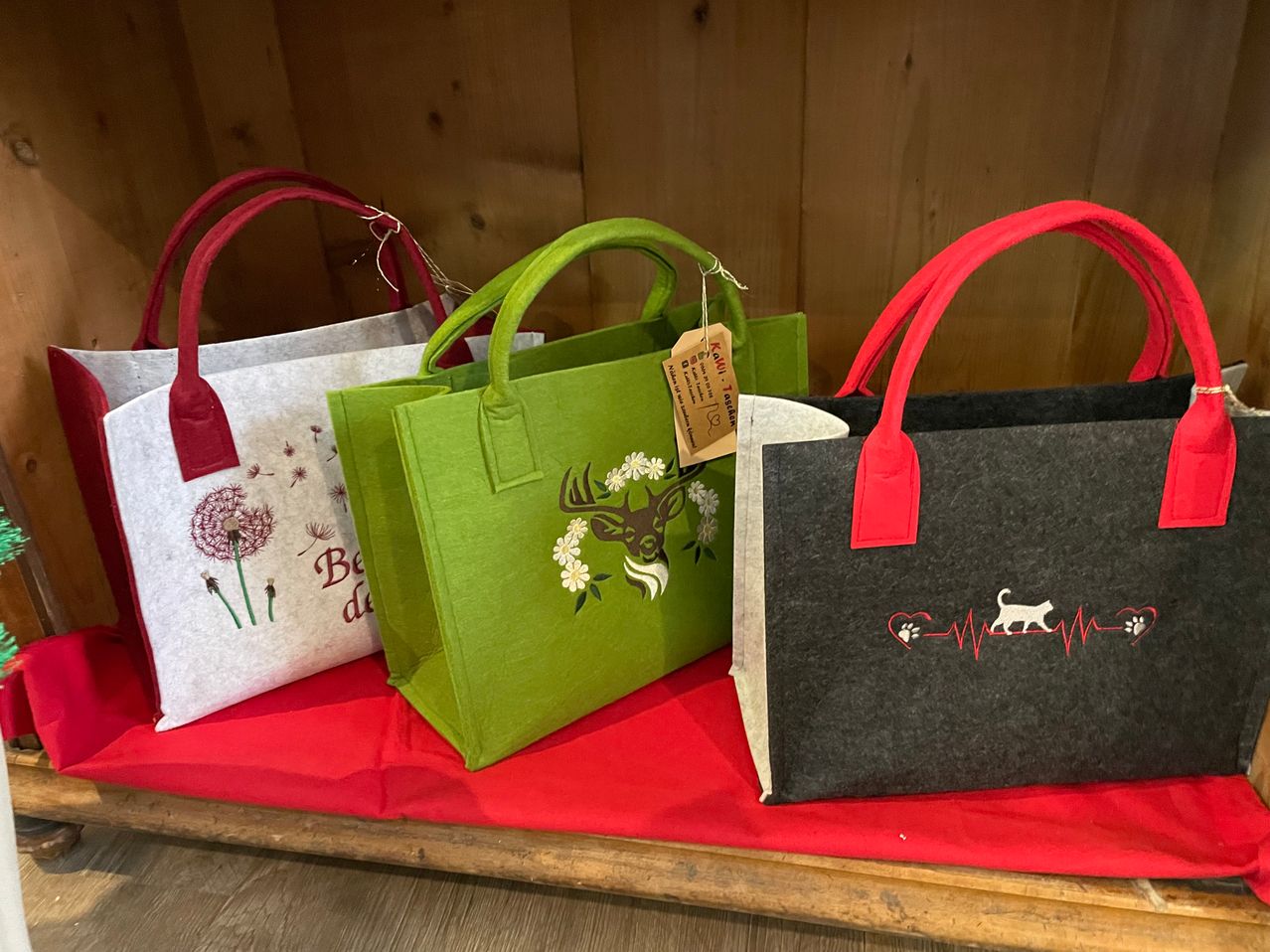 Vier Taschen mit Henkeln sind auf einem roten Tuch ausgestellt. Eine weiße Tasche hat Blumendrucke. Eine grüne Tasche hat ein Hirschdesign. Eine graue Tasche hat ein Katzendesign. Ein brauner Anhänger ist an der grünen Tasche befestigt.