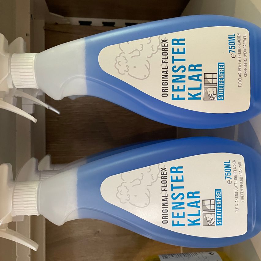 Zwei Flaschen Fenster Klar Glas- und Oberflächenreiniger, jeweils 750 ml, gestapelt auf einer Holzoberfläche. Die Flaschen haben ein blau-weißes Etikett mit einer Wolkenillustration und Produktinformationen.