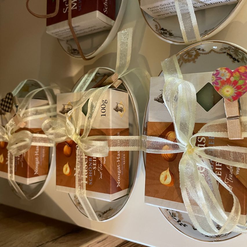 Mehrere Schachteln mit Nougats, jeweils in ein weißes Band gewickelt, sind auf einem weißen Regal ausgestellt. Die Schachteln sind mit Bildern von Haselnüssen und Herzen verziert.