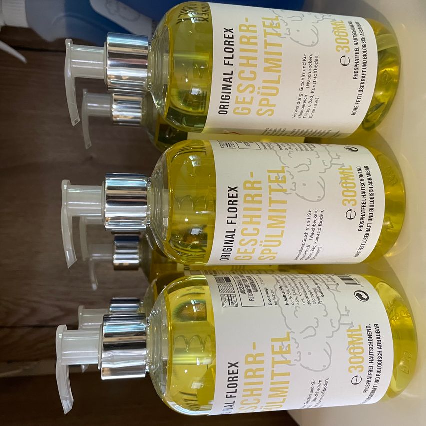 Drei Flaschen mit gelber Flüssigkeit, gekennzeichnet mit 'Original Florex,' sind in einer Reihe gestapelt. Die Etiketten zeigen Illustrationen von Schafen und den Produktnamen 'Geschirr-Spülmittel.' Die Flaschen stehen auf einer Holzoberfläche.