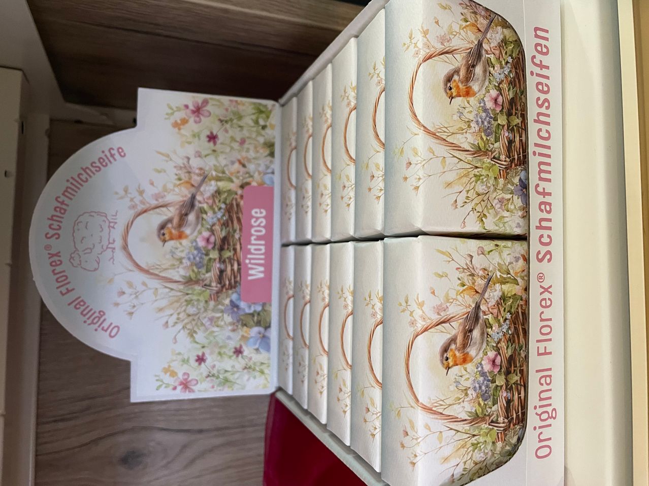 Ein Set von sechs Seifenstücken in einer Schachtel mit Blumen- und Vogeldesign. Das Etikett sagt 'Wildrose' und die Schachtel trägt den Titel 'Original Florex Schafmilchseife'. Die Schachtel ist auf einer Holzoberfläche geöffnet.