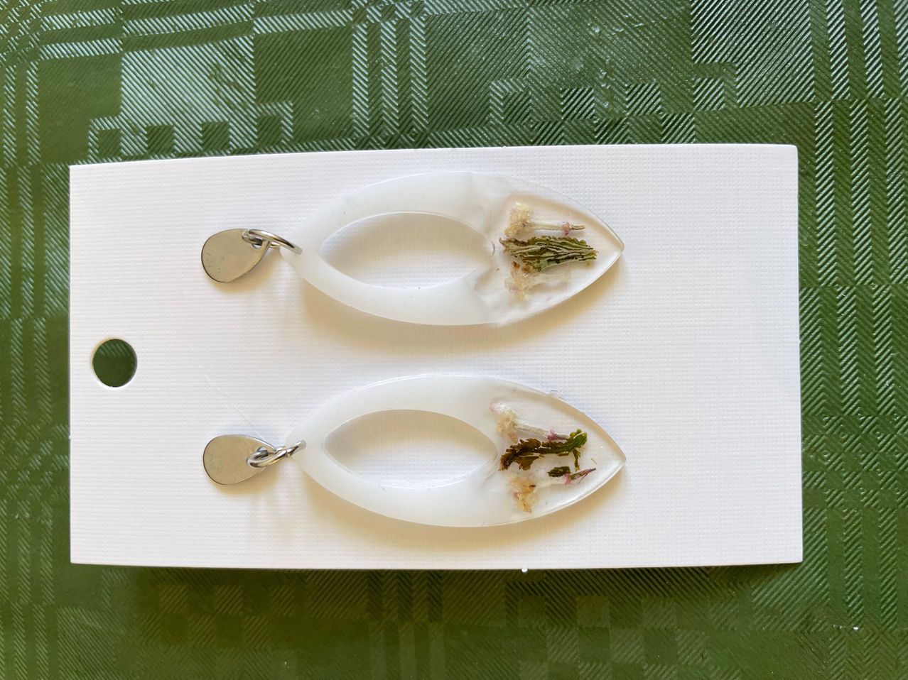 Two white earrings with oval shapes and dried flowers encased in clear resin. The earrings hang from silver hooks on a white background with a green background.