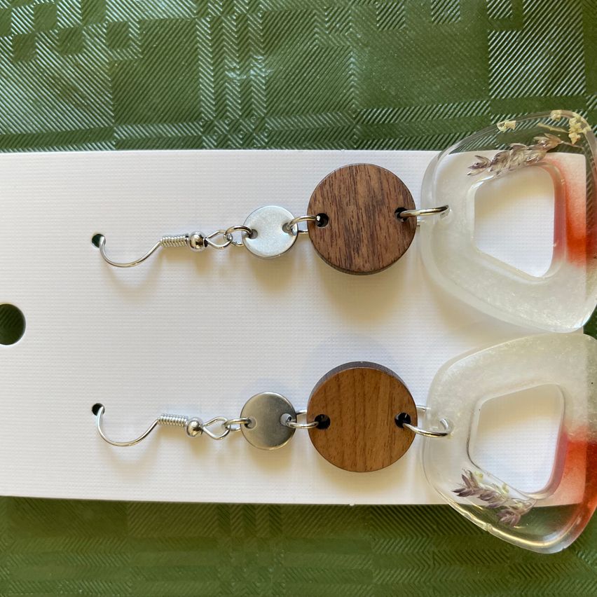 Two wooden circle earrings with a small silver bead and clear plastic drop. Each earring features a small lavender flower inside the plastic. Displayed on a white background.