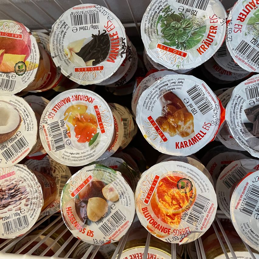 Multiple ice cream tubs of different flavors are displayed in a freezer, including Vanilla, Mandarin, Haselnuss, and Blutorange. Each tub has a barcode and a label with ingredients and nutritional information.