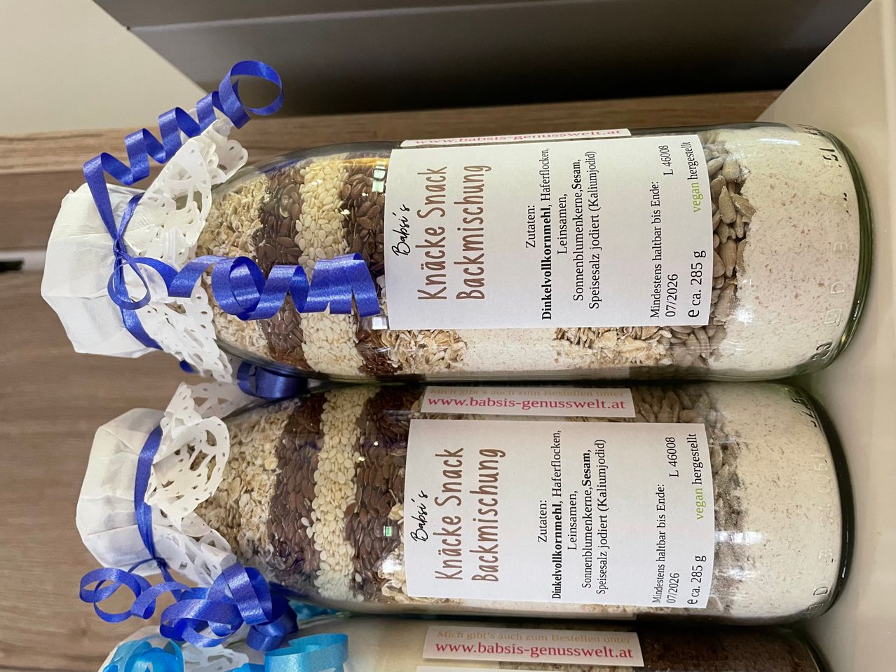 Two glass bottles with blue ribbons, filled with a mix of seeds and grains. The labels on the bottles read 'Baby's Knack-Snack' and 'Backmischung'.