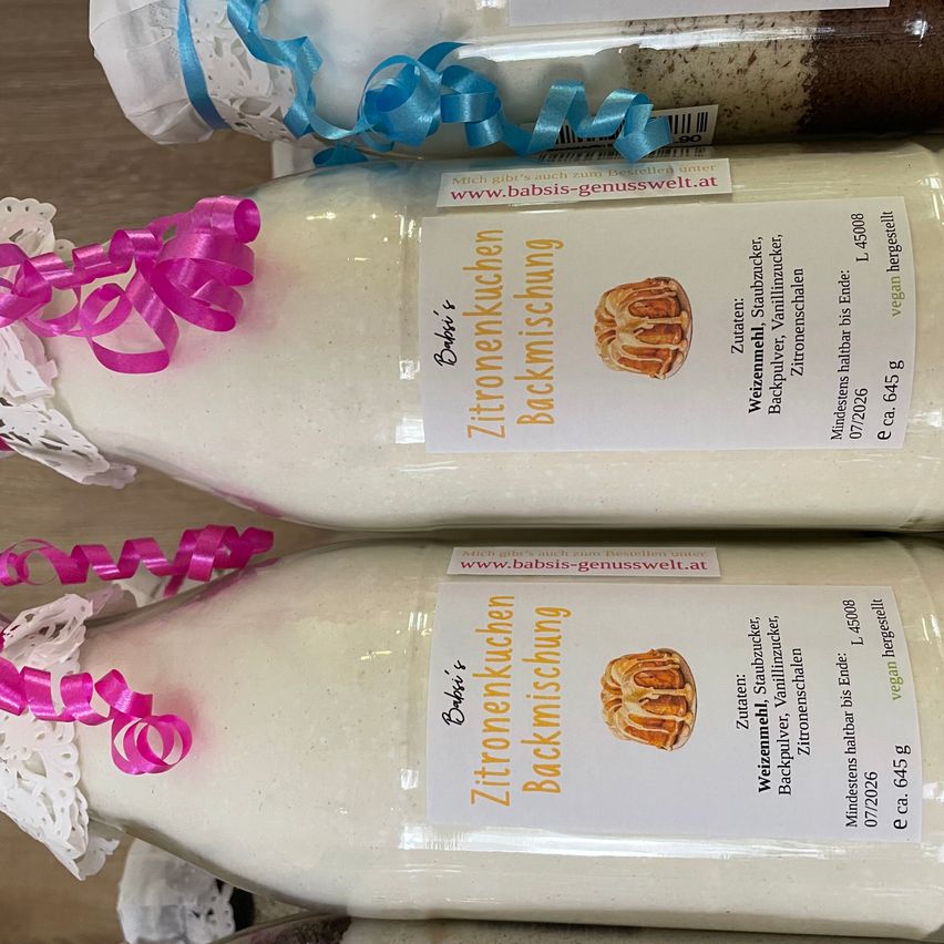 Two bottles of baking mix for cake with decorative ribbons and lace on a wooden surface. Labels include 'Zitronenkuchen Backmischung' and 'www.babsis-genusswelt.at'.