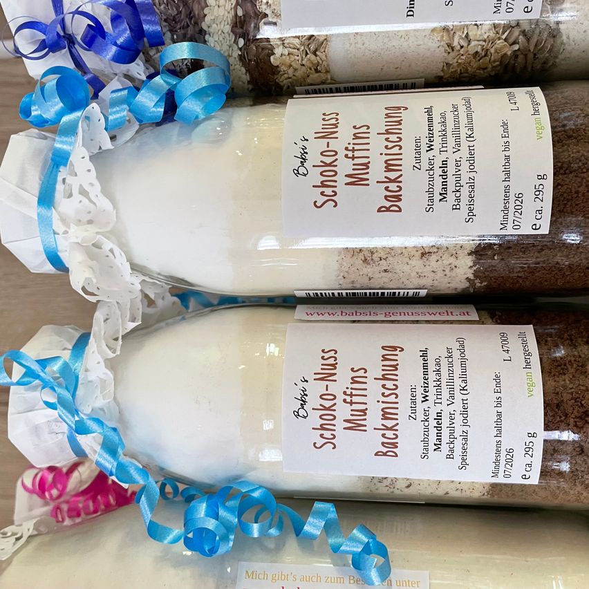 Four bottles of Baby Schoko-Nuss Muffins Backmischung, wrapped in white and blue paper, are stacked on top of each other. Each bottle contains a mix for making chocolate and nut muffins.