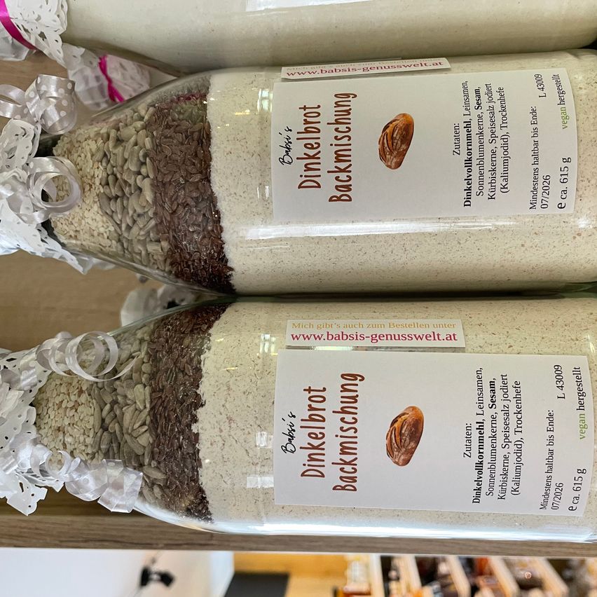 Three glass bottles with white and brown powders, tied with white ribbons, stand on a wooden shelf. Each bottle has a label with 'Dinkelbrot Backmischung' and a nut illustration.