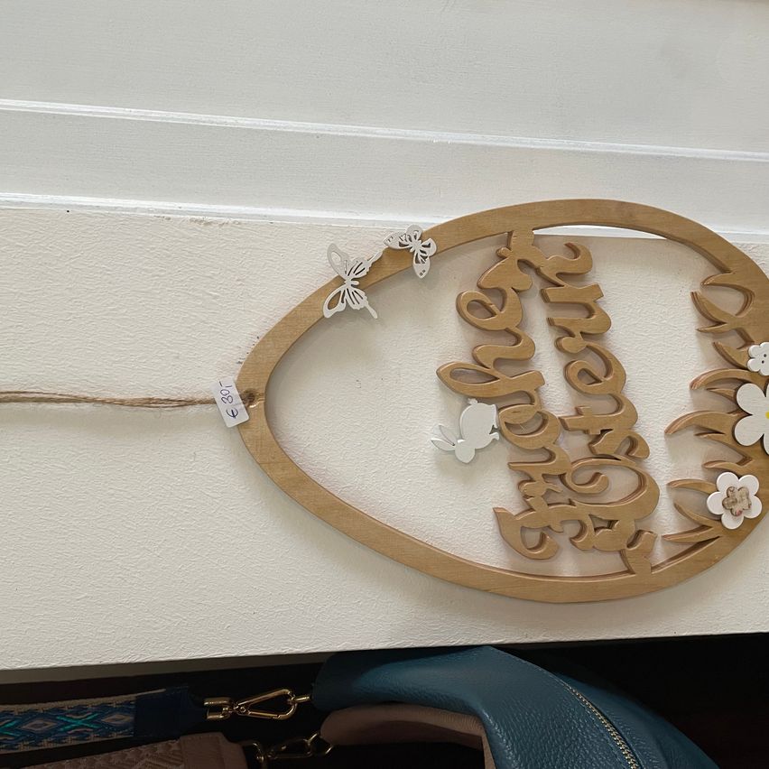 Eine hölzerne Osterdekoration mit den Worten Happy Easter und weißen Schmetterlingen und Blumen, die an einer weißen Wand befestigt sind. Darunter ist eine blaue Handtasche zu sehen.