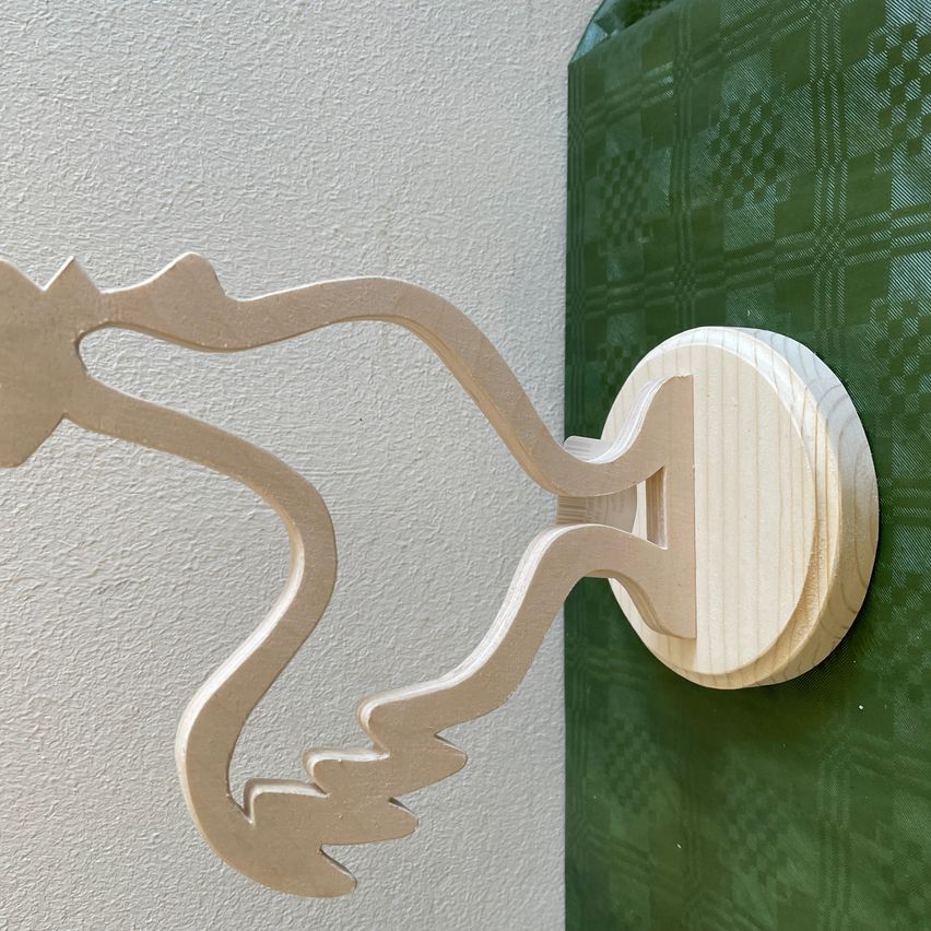 Ein hölzerner Wandhaken mit schwan-ähnlichem Design an einer weißen Wand, neben einem grünen Stoff.