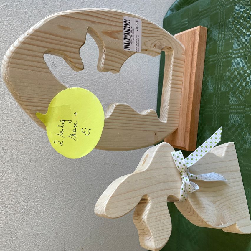 Zwei Holzskulpturen von Tieren sind an einer weißen Wand ausgestellt. Eine hat einen gelben Zettel mit handgeschriebenem Text. Eine andere hat ein Band mit grünen Punkten.