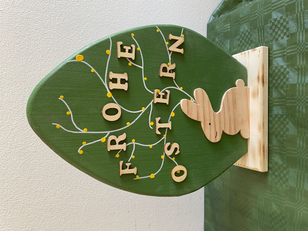 Ein holzfarbenes Schild in Form eines Blattes zeigt den Text 'FROHEN OSTERN' in weißen Buchstaben. Ein holzfarbener Hase mit gelben Punkten ist am Schild befestigt. Das Schild ist an einer Wand montiert.