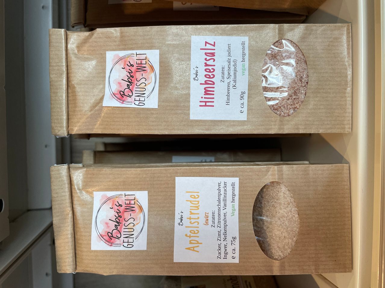 Zwei braune Kartons, die aufeinandergestapelt sind. Jeder Karton hat ein Etikett mit einem rot-weißen Design, der obere sagt 'Himbersalz' und der untere sagt 'Apfelstrudel'. Beide Etiketten enthalten den Text 'Baby's GENUSS-WELT'.
