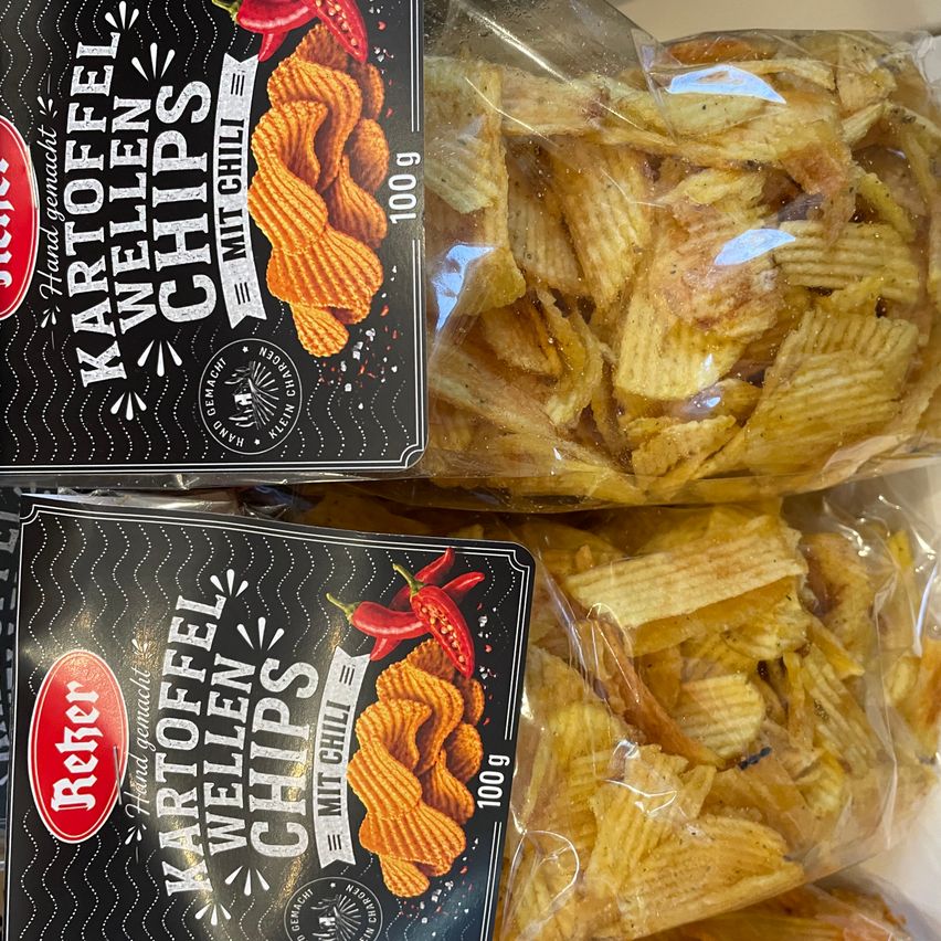 Zwei Tüten mit welligen Kartoffelchips mit milder Chili-Geschmack sind übereinander gestapelt. Die Tüten haben einen schwarzen Hintergrund mit weißem Text und einer roten Chili-Illustration.