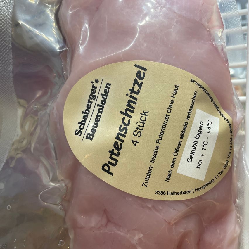 Ein Stück rohes Hähnchenbrustfleisch mit einem Etikett, das 'Putenschniitzel' von 'Schaberger's Bauernladen' liest. Das Etikett zeigt an, dass es frisch ist, ohne Konservierungsmittel, und sollte bei einer Temperatur zwischen 1°C und 4°C gelagert werden.