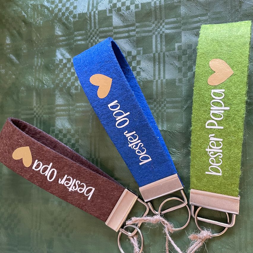 Three keychains with key rings, one blue, one green, and one brown. Each features a heart and the word 'bester' or 'papa' in different languages. They rest on a green patterned cloth.
