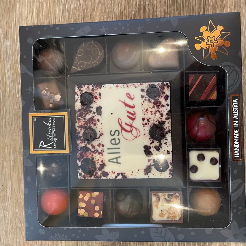 A box of assorted chocolates with a central piece labeled 'Alles Gute', surrounded by various chocolates and candies, including a red-covered fruit and a chocolate-coated nut.