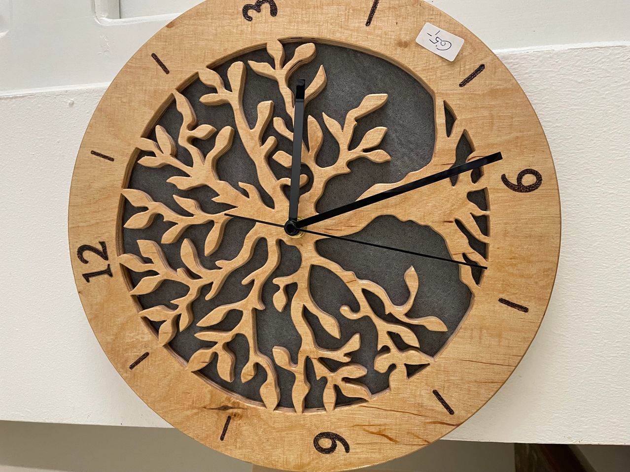 A wooden round clock with a gray tree design on its face. Numbers 1 to 12 are marked in black. A price tag with -50 is attached to the upper right.