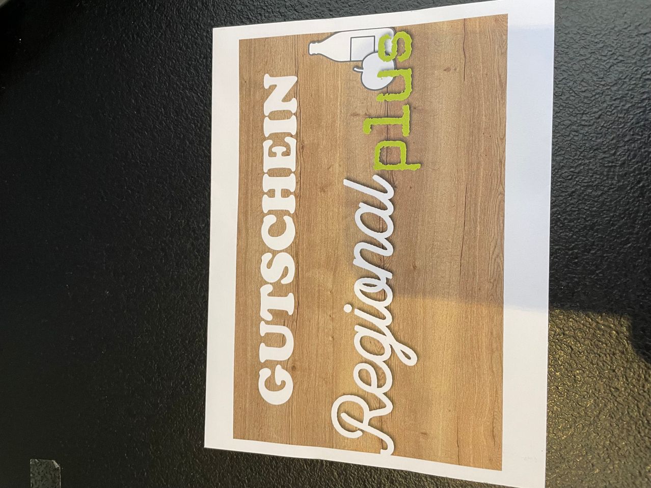 Ein hölzerne Tafel mit weißem Text, der 'Regional' und 'Gutschein' auf dunklem Hintergrund liest. Das Wort 'Gutschein' ist in fetten weißen Buchstaben, während 'Regional' in kursiver Schrift mit einem grünen Apfel-Grafik geschrieben ist.