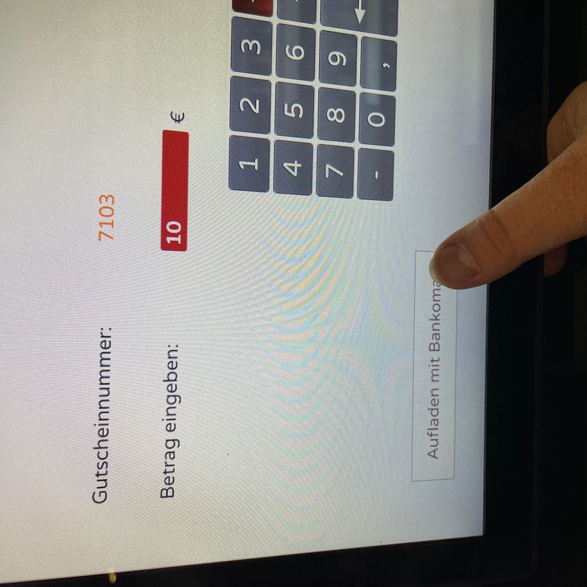 Ein Screenshot zeigt ein Gerät mit einer digitalen Nummerntastatur, '7103', '10' und einer Schaltfläche mit der Aufschrift 'Aufladen mit Bankkonto'. Ein Finger zeigt auf eine hervorgehobene Zahl '10'.