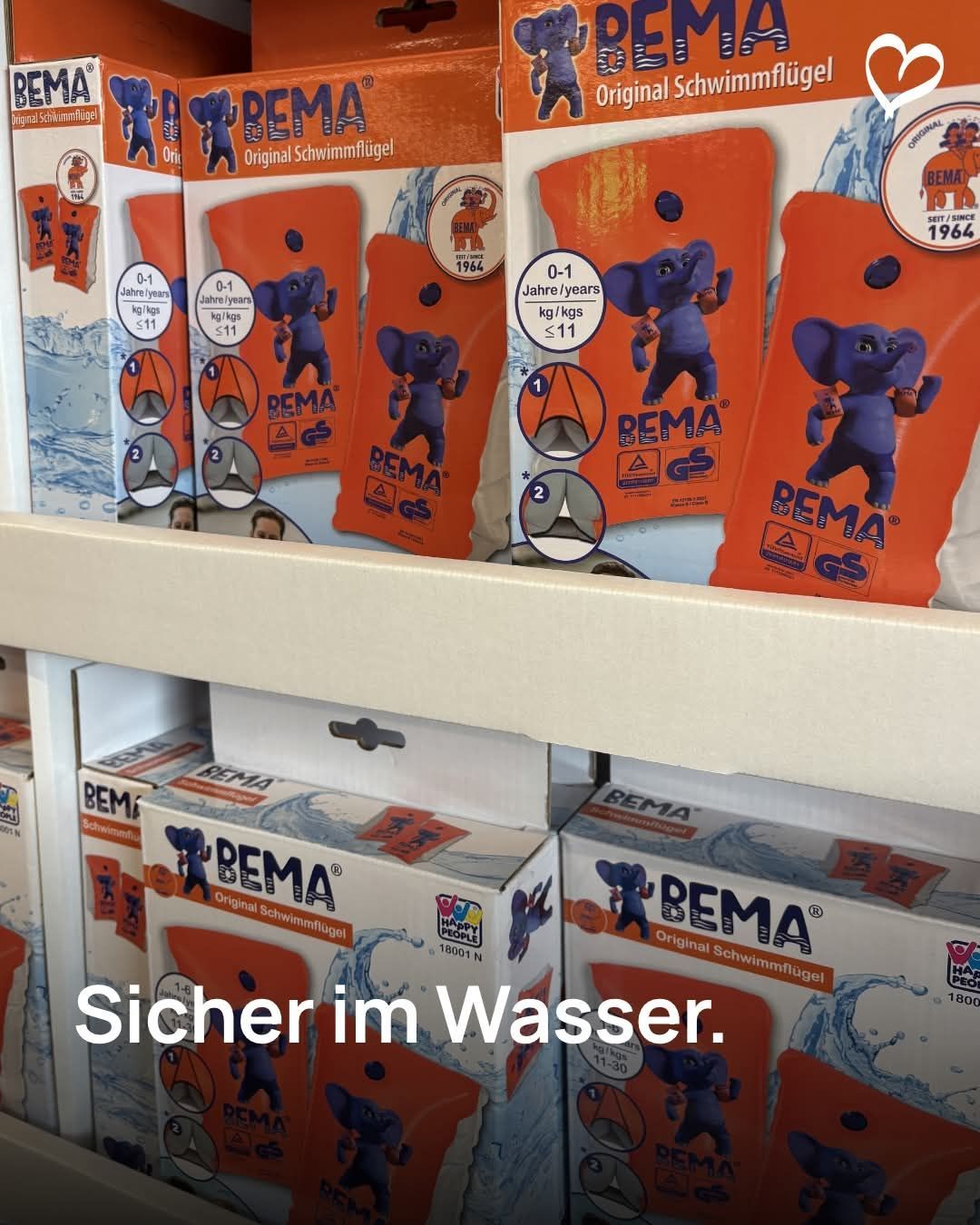 Regal mit mehreren Kartons von BEAM-Schwimmhilfen für Kinder im Alter von 0-1 Jahr. Die Kartons sind orange und weiß, mit einem blauen Elefanten-Design. Jeder Karton ist mit Gewicht und Alterseignung beschriftet. Am unteren Rand ist der Text 'Sicher im Wasser' sichtbar.