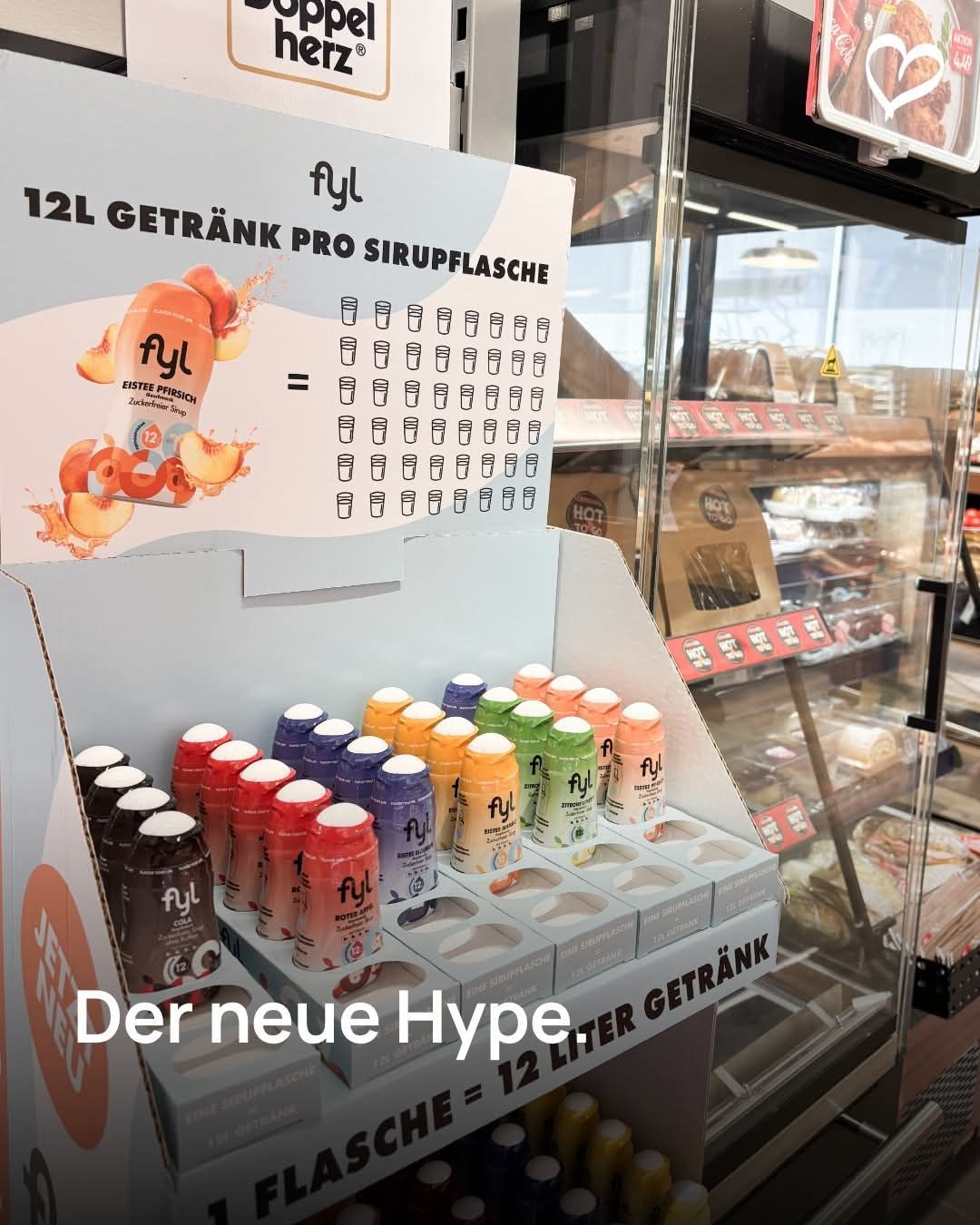Ein Display-Schrank zeigt bunte Getränkeflaschen mit der Aufschrift 'Fyl'. Der Schrank enthält 12L Sirup-Getränk und das Schild sagt 'Der neue Hype' und '12 LITER GETRÄNK'. Dahinter befindet sich ein Glasschrank mit verschiedenen Lebensmitteln.