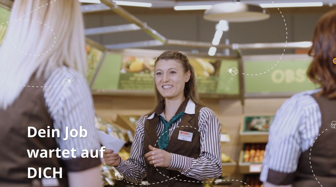 Eine lächelnde Frau in einem braunen Weste mit einer grünen Krawatte steht in einem Supermarkt, hält ein Stück Papier in der Hand. Ein Mann steht neben ihr.