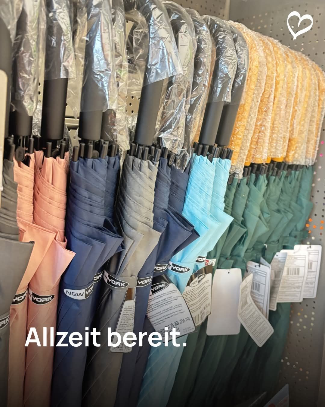 Eine Reihe von Regenschirmen, zusammengefaltet und in Plastik eingewickelt, auf einem Ladenregal ausgestellt. Die Regenschirme kommen in verschiedenen Farben und haben Etiketten mit Produktinformationen. Der deutsche Ausdruck 'Allzeit bereit' ist unten angezeigt.