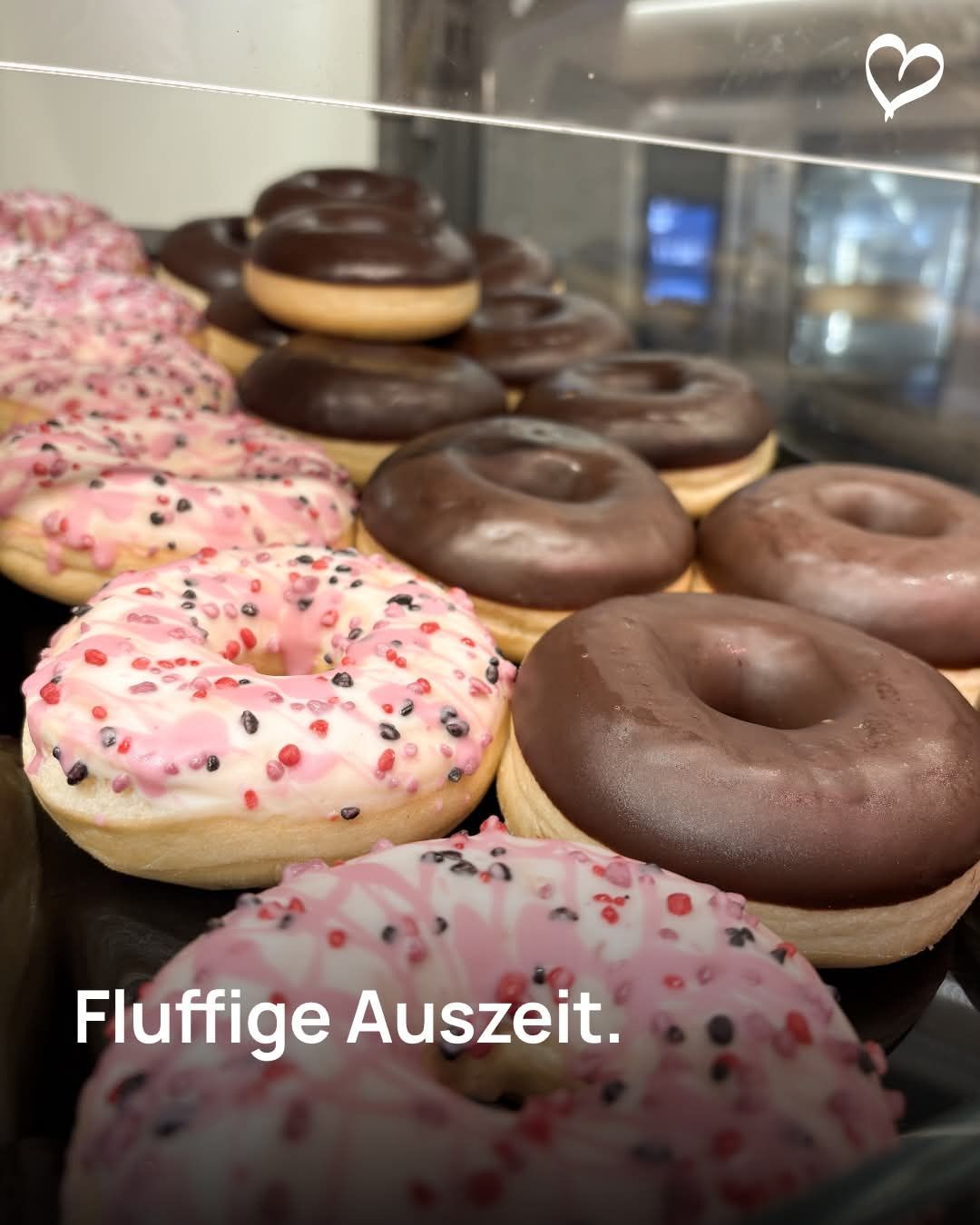 Eine Vitrine mit einer Auswahl an Donuts, darunter schokoladenglasierte, pinkfarbene mit Streuseln und mehr. Die Vitrine hat einen Glasdeckel.