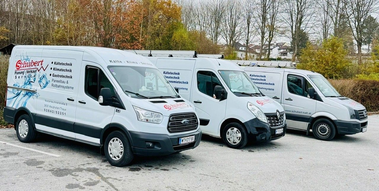 Drei weiße Lieferwagen stehen in einer Reihe auf einer Straße. Die Lieferwagen haben die Aufschriften 'Kalte Klima Service' und 'Schubert' an ihren Seiten. Dahinter stehen Bäume und Häuser.