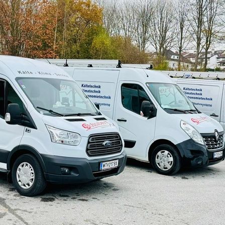 Drei weiße Lieferwagen stehen in einer Reihe auf einer Straße. Die Lieferwagen haben die Aufschriften 'Kalte Klima Service' und 'Schubert' an ihren Seiten. Dahinter stehen Bäume und Häuser.