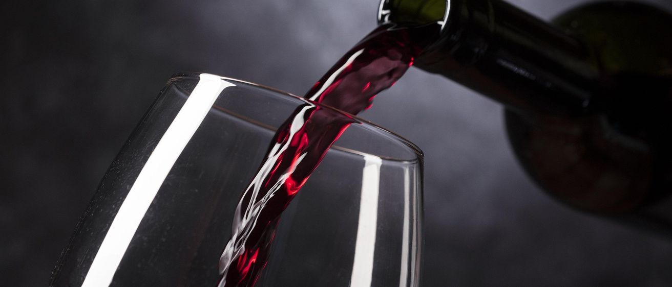 Eine dunkle Flasche gießt roten Wein in ein Glas auf schwarzem Hintergrund. Der Wein fließt in einem dünnen Strahl.