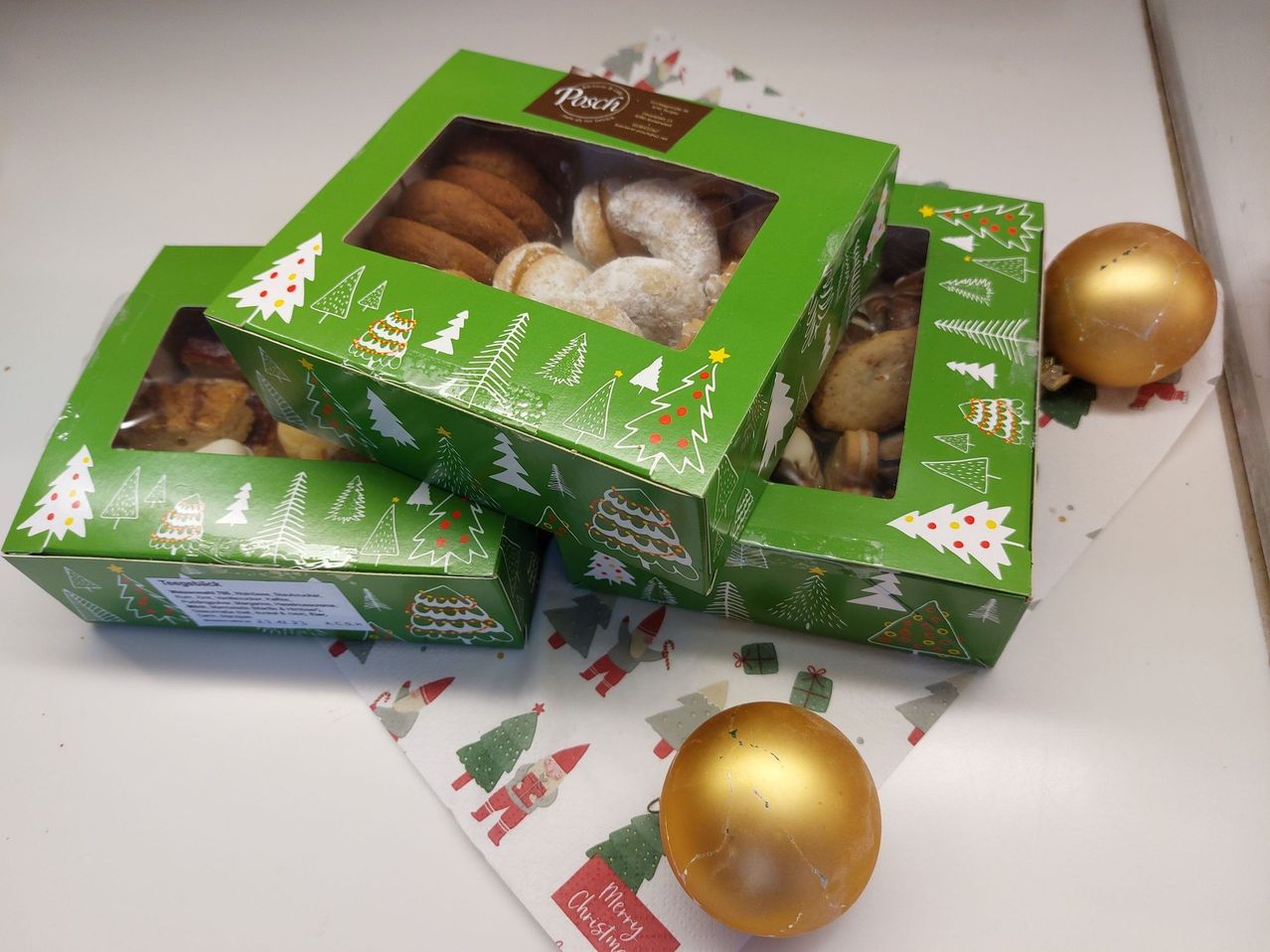Drei grüne Schachteln mit Weihnachtsbäumen und einem Fenster, das Kekse zeigt. Ein goldener Weihnachtsball liegt in der Nähe.