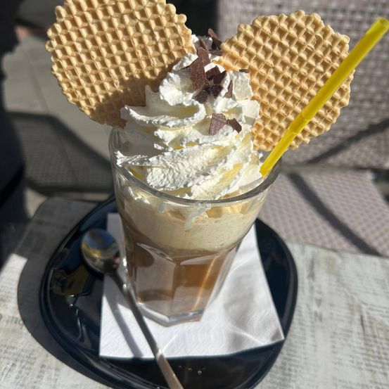 Eine Kaffee-Eiscreme mit Schlagsahne und Schokoladenwaffeln darauf, serviert in einem Glas mit einem Strohhalm, auf einem schwarzen Teller.