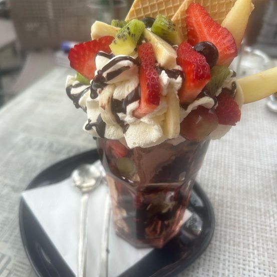 Ein Dessert in einem Glas mit verschiedenen Früchten und Schlagsahne auf einem Teller mit einem Löffel.