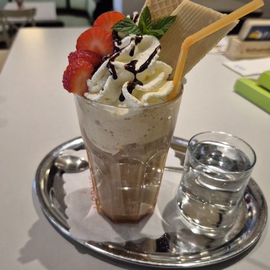 Ein Milchshake mit Schlagsahne, Schokoladensauce, Erdbeeren, Minzblättern und Eistütenstücken steht auf einem silbernen Tablett.