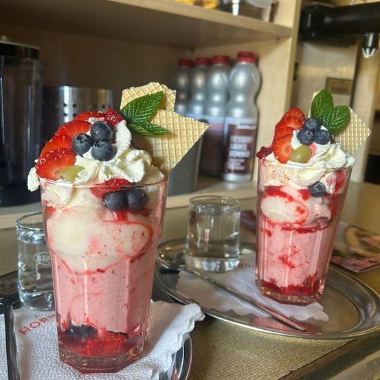 Zwei Eisbecher mit Erdbeeren, Blaubeeren, Schlagsahne und Waffeln auf einem Tisch mit einem silbernen Teller und einem kleinen Glas Wasser.