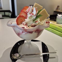 Ein Eisbecher mit Erdbeereis, Schlagsahne und Erdbeerscheiben mit einem Löffel auf einem schwarzen Teller.