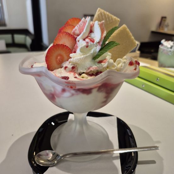 Ein Eisbecher mit Erdbeereis, Schlagsahne und Erdbeerscheiben mit einem Löffel auf einem schwarzen Teller.