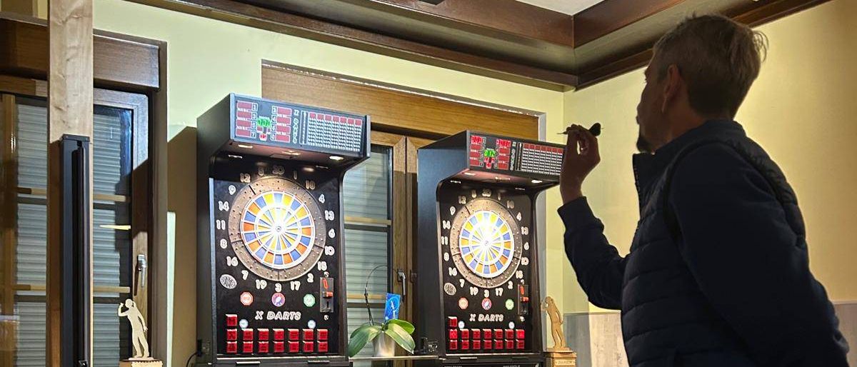 Ein Mann in einer blauen Jacke wirft Darts auf ein Dartboard in einem Spielzimmer mit Dart-Kabinetten und weißen Schränken.