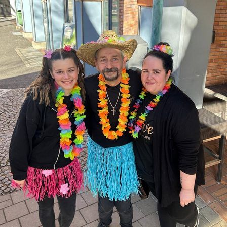 Ein Mann und zwei Frauen posieren für ein Foto in bunten Leis und Hüten. Sie stehen auf einem Ziegelweg mit einem Ziegelgebäude und einer Glastür im Hintergrund.