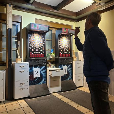 Ein Mann steht vor zwei Dart-Automaten, der eine mit '2' und der andere mit '1' beschriftet. Er hält einen Dart und zielt auf das Brett.